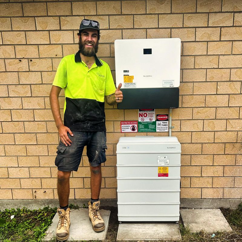 An installer giving a thumbs up beside a newly installed inverter and battery on a brick wall