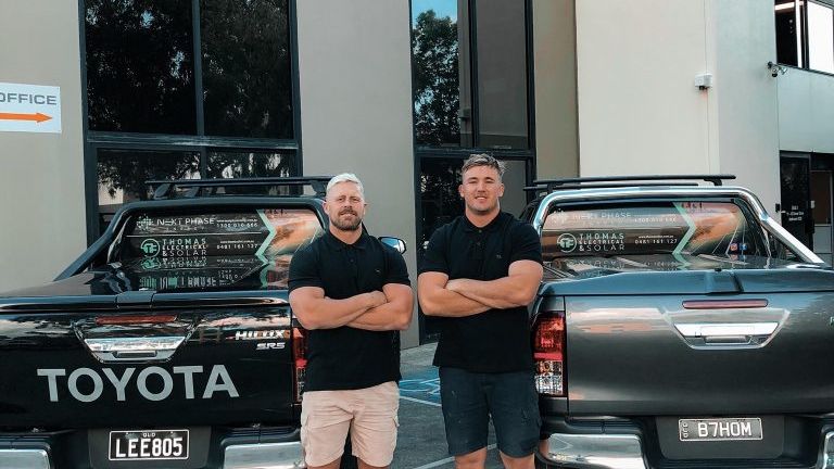 Two Thomas Electrical and Solar team members standing in front of their work utes outside the workshop